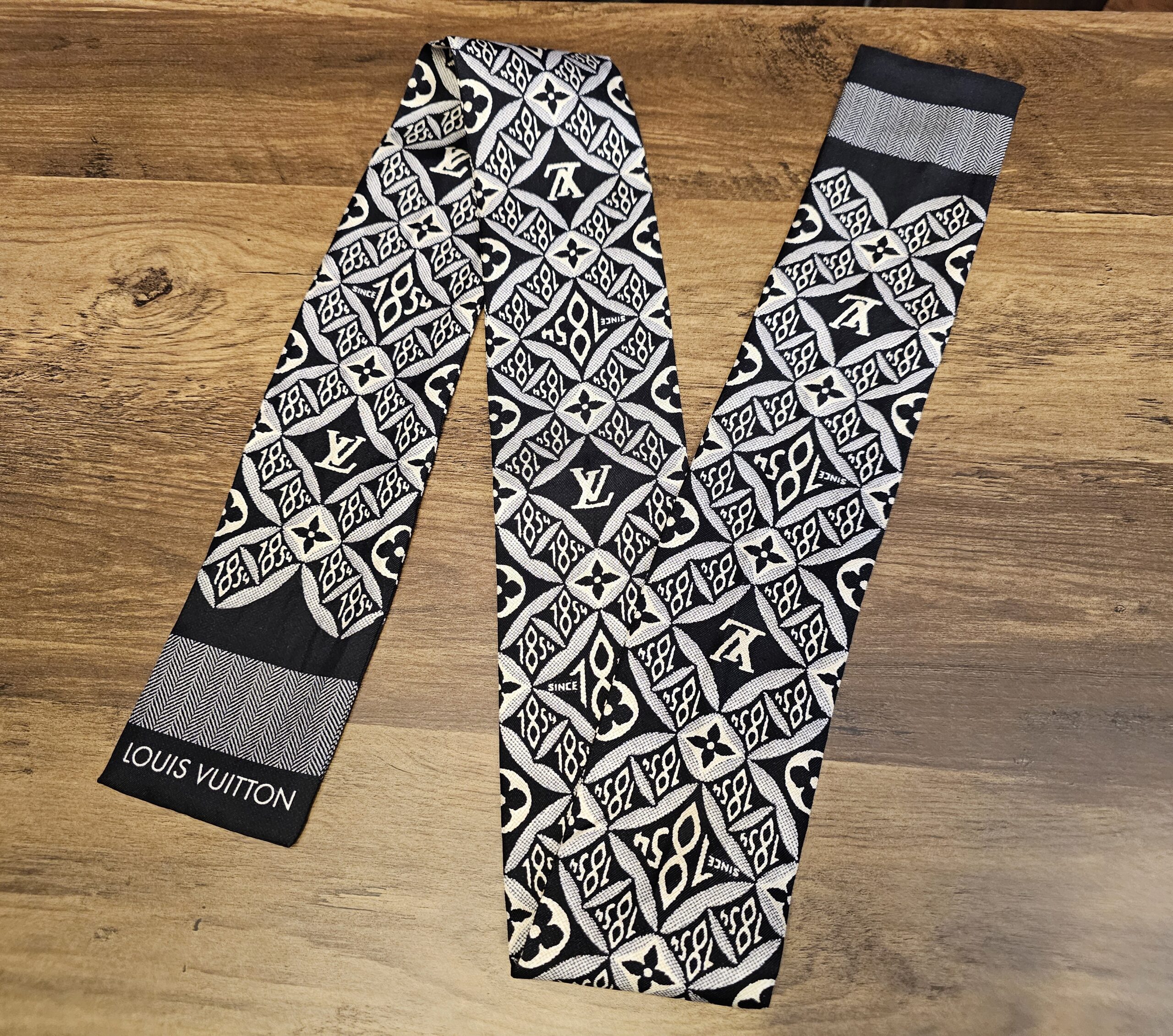 Louis Vuitton Silk Since 1854 Bandeau Black Cream Gray laid out on a wood table with the Louis Vuitton name on the end